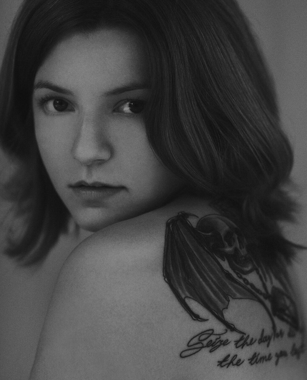 Black and white Portrait of a girl with tattoo on her shoulder; death's head with wings; Seize the day or die regretting the time you lost